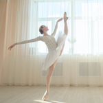 Image of a ballerina in front of a big window with sheer curtains. She is reaching up with her leg and arm