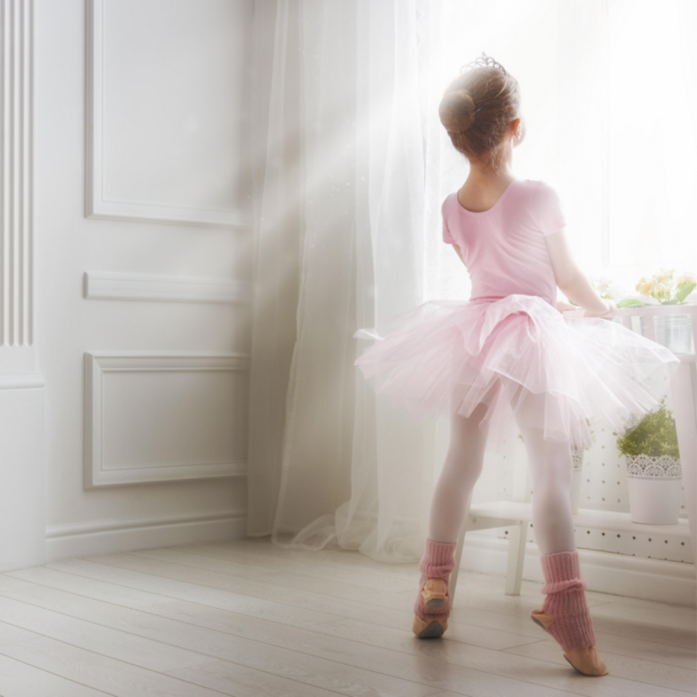 Little girl in her ballerina outfit standing on her toes and wearing a crown, looking out a bright window