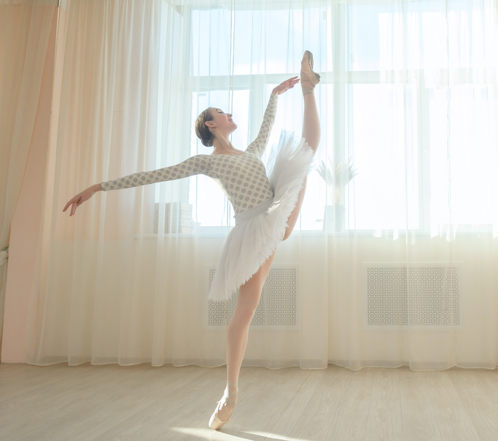 Ballerina with her leg extended and arms out infront of a huge window with sheer curtains
