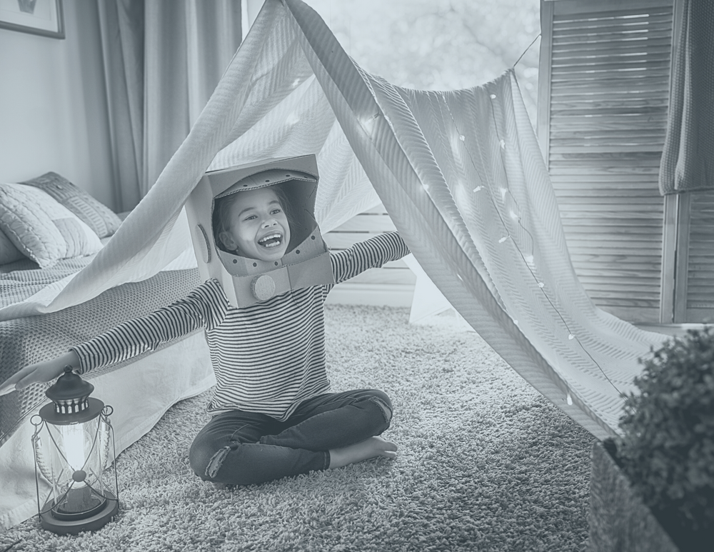 Image of a kid in her fort in her room with twinkle lights and an astronaut helmet made from cardboard