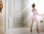 Young girl with ballet outfit on standing on her toes and looking out a bright window. She is wearing a crown