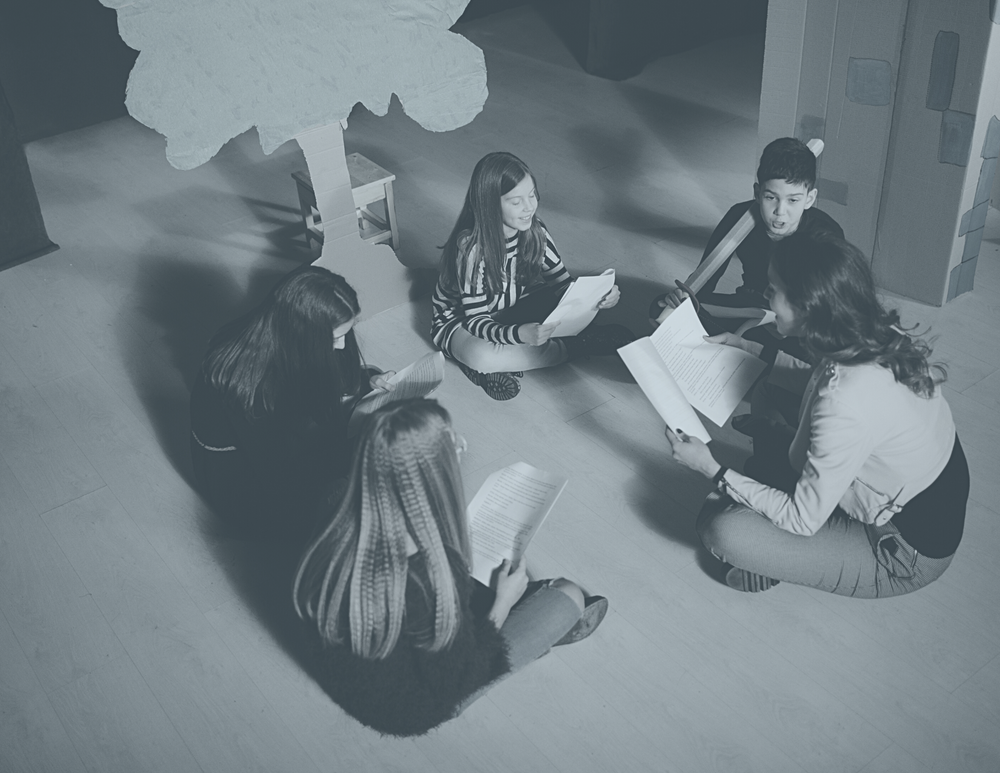 Image of kids sitting around a circle with theatre props around them, they are holding a script and running lines