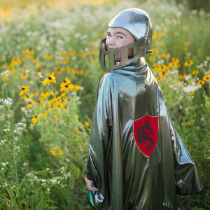 
                  
                     Great Pretenders Costume of a boy in a field wearing a silver shinny cape and a red and black dragon symbol on it.
                  
                