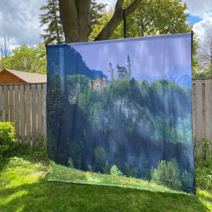 
                  
                    Showing the full backdrop set up under a tree in a backyard. It's displayed on it's stand and features the image of a castle and mountains
                  
                